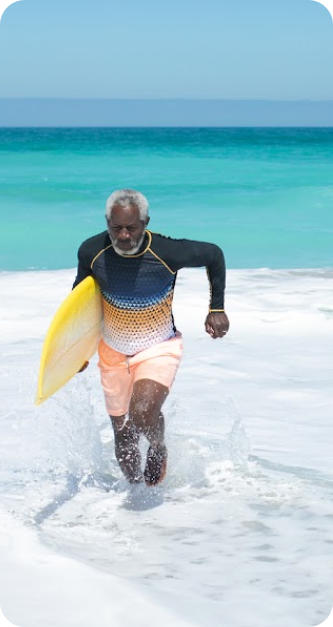 Photo of a senior man with a surfboard under his arm running through the shallow surf of a tropical beach