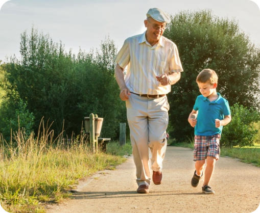 A senior man is jogging down an outdoor path with his school-age grandson