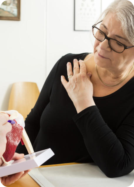 A woman holds her right shoulder with her left hand as she looks at a model of a shoulder. The doctor is pointing to something on the shoulder model.