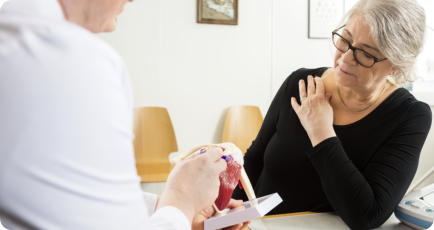 A woman holds her right shoulder with her left hand as she looks at a model of a shoulder. The doctor is pointing to something on the shoulder model.