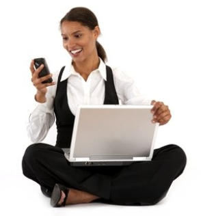 Photo of a young woman with a white background. She is sitting cross-legged with her computer on her lap and is smiling at her phone.