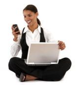 Photo of a young woman with a white background. She is sitting cross-legged with her computer on her lap and is smiling at her phone.